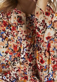 Blusa floral con patrones en rojo, naranja, azul y negro, con frente plisado, botones blancos y mangas largas abullonadas.