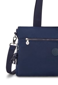 Navy blue nylon bag with a zippered front pocket, adjustable strap, and a rubber charm. Features a round logo patch and subtle stitching details.