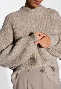 Beige knitted sweater with a high neckline, ribbed texture, and loose sleeves. The fabric appears soft and cozy, with subtle detailing.