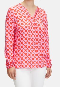 Femme portant un blouse rouge à manches longues avec un motif de feuilles géométriques roses et un col en V, assortie à un pantalon blanc, vue de la poitrine aux hanches.