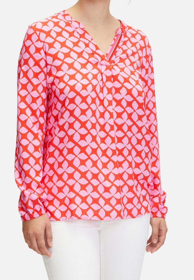 Femme portant un blouse rouge à manches longues avec un motif de feuilles géométriques roses et un col en V, assortie à un pantalon blanc, vue de la poitrine aux hanches.