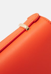 Detail oranžové kožené kabelky se hladkým povrchem, šitými okraji a zlatým uzávěrem.