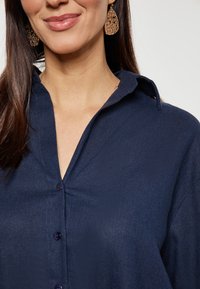 Camicia blu scuro con colletto, caratterizzata da una texture sottile e bottoni scuri. Abbinata a orecchini intricati a forma di goccia.