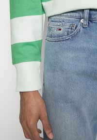 Camisa de manga larga a rayas verdes y blancas con puños acanalados, combinada con jeans de denim azul claro que presentan una pequeña etiqueta de logo en el bolsillo.