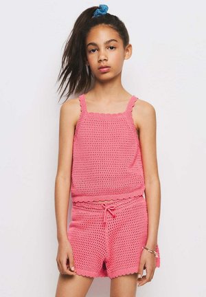 Jeune fille avec une haute queue de cheval portant un débardeur rose en crochet et un short assorti à cordon sur un fond uni.