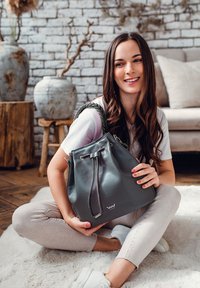 Šedá kožená kabela ve tvaru vědra s pleteným popruhem, stahovací šňůrkou a logem. Model sedí na měkkém koberci, v neformálním oblečení a bílých teniskách.