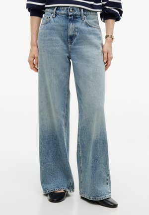 Person iført højtaljede, vidde lyseblå jeans kombineret med sorte spidse sko og en stribet langærmet top, stående med afslappede hænder.