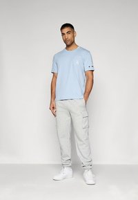 Gaiši zila kokvilnas T-krekls ar kabatu, kopā ar pelēkām sporta biksēm ar sānu kabatām. Aksesuāri ietver baltus sporta apavus.