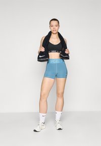 Soutien-gorge de sport noir Nike, short taille haute bleu clair avec un logo subtil, chaussettes blanches de sport, et chaussures de sport blanches, tenant une serviette noire.