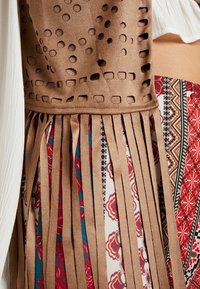 Braunes Wildleder mit ausgeschnittenen Mustern und langen Fransen, übereinandergelegt auf einem gemusterten Stoff mit floralen und geometrischen Designs.