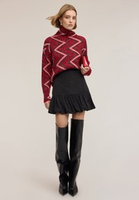 Suéter de punto con patrón de zigzag en rojo y rosa, minifalda negra con volantes y botas de cuero negras hasta el muslo. Sosteniendo un pequeño bolso rojo.