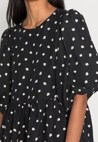 Musta mekko, jossa on tekstuuri ja valkoisia kukkakuoseja, puffatut hihat ja kerrottu vyötärö. Pyöreä pääntie ja pehmeä, virtaava siluetti.