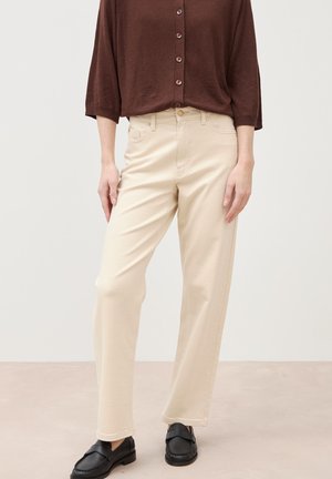 Personne portant un pantalon beige taille haute à jambes droites, un haut marron boutonné avec manches trois-quarts, et des chaussures noires à enfiler sur un fond neutre.