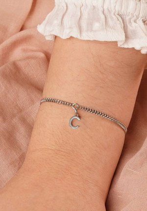 Silver chain bracelet with a dangling letter "C" charm worn on a wrist against a soft pink fabric background.