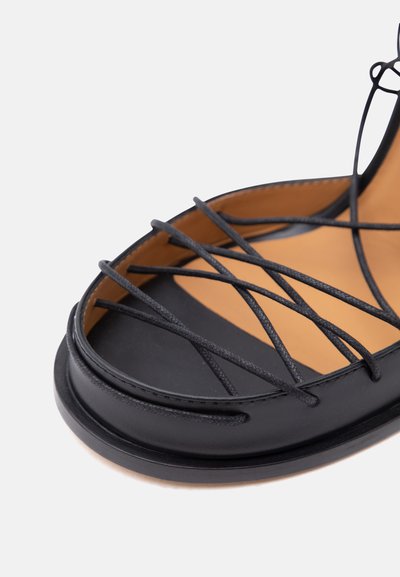 Μαύρο επίπεδο σανδάλι με πολλαπλούς λεπτούς σταυρωτούς λουράκια πάνω σε ταμπά πάτο και στρογγυλεμένη μύτη σε λευκό φόντο.