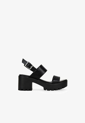 Sandalias de plataforma de cuero negro con un tacón robusto, correa en el tobillo ajustable y diseño cruzado en la parte superior. Suela texturizada para un mejor agarre.