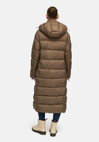 Braune Pufferjacke mit Kapuze, lang geschnitten, gestepptes Design und sichtbare Reißverschlüsse an den Ärmeln, kombiniert mit beigen Schnürstiefeln.
