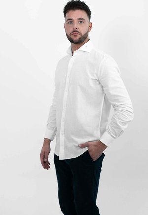 Man met donker krullend haar en baard, draagt een wit overhemd met knopen en donkere broek, staat met één hand in zijn zak.