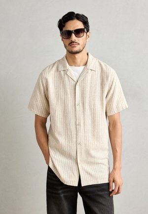 Camicia a maniche corte con colletto in beige chiaro con righe verticali in tonalità scure; il tessuto sembra texturizzato. Abbinata a jeans scuri.