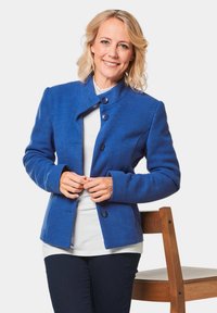 Blaue Wollmischung-Jacke mit hohem Kragen, Knopfverschluss und langen Ärmeln. Getragen über einem weißen Oberteil und dunklen Jeans, mit einem einfachen Holstuhl im Hintergrund.