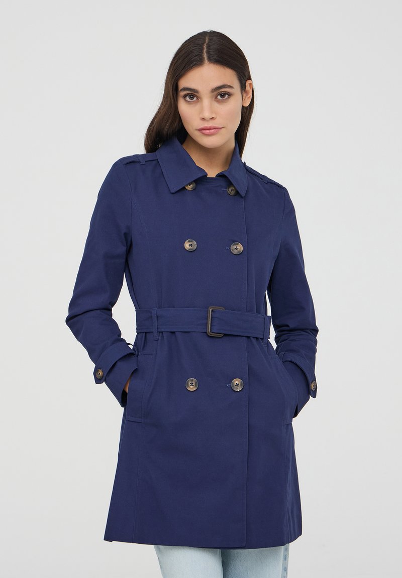 Cappotto trench blu navy con design doppiopetto, vita con cinturino e dettagli con bottoni. Presenta un colletto e tasche laterali.