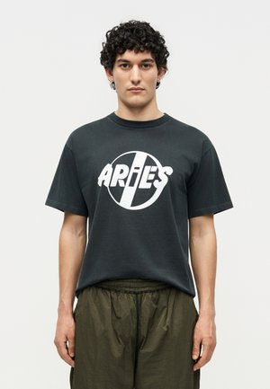Mann mit lockigem Haar, der ein schwarzes T-Shirt mit Widder-Logo und olivgrünen Hosen trägt, steht vor einem schlichten hellen Hintergrund.