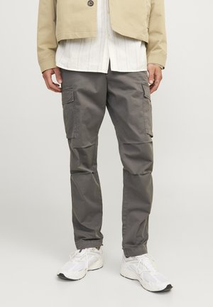 Pantaloni cargo grigi con molte tasche laterali, design affusolato e una texture morbida e opaca, abbinati a sneakers bianche e a una giacca beige.
