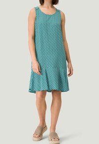 Ærmeløs turkis kjole med geometrisk mønster, A-formet silhuet, letvægtsstof og en udvidet kant. Båret med beige slip-on sandaler.