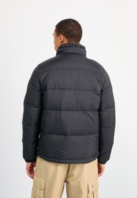 Giacca imbottita nera con alto colletto, tessuto testurizzato, chiusura con zip e polsini elasticizzati. Indossata sopra pantaloni cargo beige.
