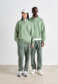 Green zip-up sweatshirts and patterned trousers feature a geometric design in shades of green. White sneakers complete the outfits.