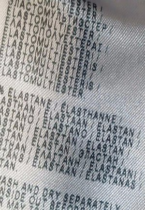 Návod na údržbu odevu uvádzajúci zloženie látky, prevažne polyester a elastan, v niekoľkých jazykoch na bielom textúrovanom materiáli.