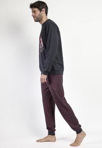 Camisa negra de manga larga con diseño gráfico, combinada con pantalones rojos estampados con puños elásticos. Modelo de pie descalzo.