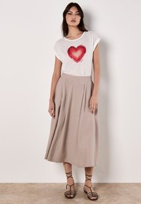 T-shirt blanc avec un graphique de cœur rouge, associé à une jupe midi plissée beige. Le mannequin porte des sandales noires à lanières. Textures de tissu douces.