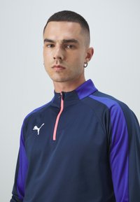 Tamsiai mėlynas sportinis marškinėlis su violetinėmis rankovėmis, turintis pusiau užtrauktuką priekyje ir rožinį užtrauktuką. Ant krūtinės yra baltas Puma logotipas. Lygi tekstūra.