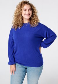 Koningsblauwe oversized trui met geribbelde manchetten en zoom, zachte textuur, ronde halslijn, gecombineerd met lichtblauwe jeans. Geaccentueerd met gelaagde kettingen.