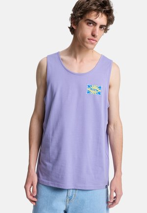 Młody mężczyzna z falującymi brązowymi włosami, ubrany w lawendową koszulkę bez rękawów z małym logo słońca i fali, w połączeniu z jasnoniebieskimi szortami dżinsowymi.