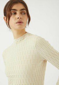 Beige texturerad långärmad topp med hög hals och ett vävt mönster, med ribbade detaljer längs sömmarna och ärmarna.