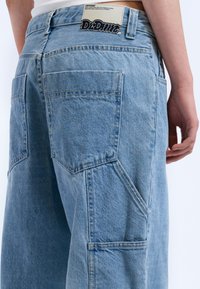 Light blue denim jeans feature a medium-rise waist, large back pockets, and a branded patch at the waistband. Textured fabric with visible stitching.