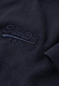Mørkeblåt stof med broderet "SuperDry"-tekst og japanske tegn i matchende tråd, som viser en del af en foldet ærme.
