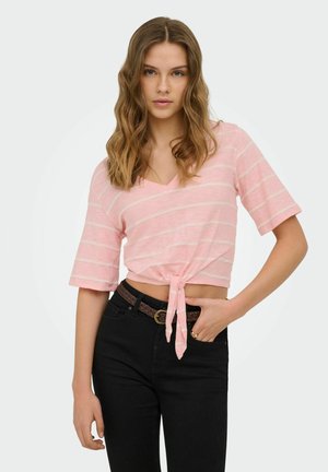 Jeune femme aux cheveux ondulés portant un crop top rayé rose noué à la taille et un pantalon noir avec une ceinture tressée marron.