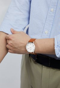 Montre à cadran blanc avec matériel en argent, bracelet en cuir marron et marquages simples en noir. Portée au poignet, assortie à une chemise à rayures bleues.