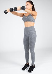 Conjunto de entrenamiento gris con un sujetador deportivo ceñido y leggings de cintura alta. La figura sostiene dos mancuernas hexagonales negras. Zapatillas deportivas negras.