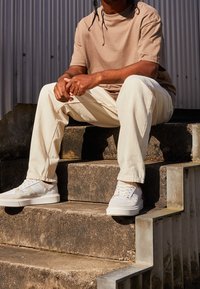 Béžové tričko s krátkým rukávem, off-white džíny a bílé tenisky s akcenty, sedící na kamenných schodech proti vlnitému pozadí.