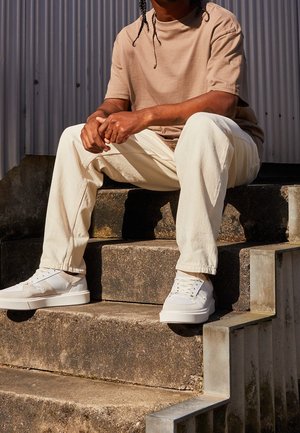 Camiseta beige de manga corta, pantalones de mezclilla color blanco roto y zapatillas blancas con acentos, sentado en escalones de piedra contra un fondo acanalado.