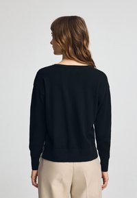 Sweater noir tricoté avec un col rond, épaules tombantes et ourlet côtelé. Porté sur un pantalon beige, mettant en valeur une coupe décontractée.