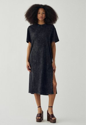 Kobieta o bujnych, kręconych włosach, ubrana w czarną, midi sukienkę na krótki rękaw z bocznym rozcięciem oraz brązowe platformy na obcasie.