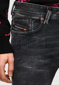 Jeans de mezclilla negra con detalles desgastados en el bolsillo delantero, una etiqueta de marca y herrajes de cobre. Primer plano de una mano agarrando la pretina.