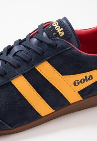 Baskets en suède bleu marine avec deux bandes jaunes, semelle en caoutchouc marron et doublure intérieure rouge. Logo Gola sur la languette et sur le côté.