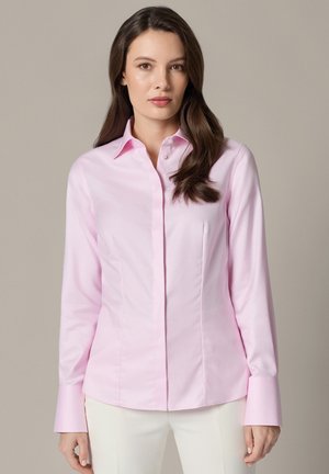 Kobieta z długimi brązowymi włosami, ubrana w jasnoróżową dopasowaną koszulę z guzikami i białe spodnie, stojąca na beżowym tle.