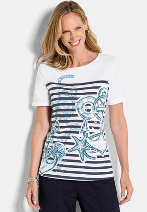 Vrouw met blond haar die lacht, gekleed in een wit shirt met korte mouwen met marineblauwe strepen en blauw nautisch thema, gecombineerd met een donkere broek.